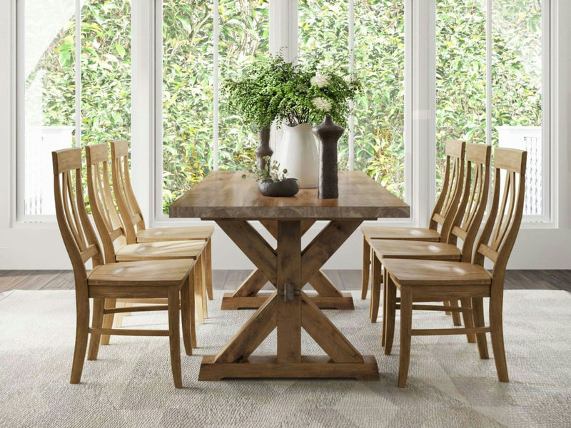 A Trestle Dining Table in Harvest Wheat from James+James, featuring an X-shaped base, is surrounded by eight matching wooden chairs. The table is adorned with vases of white flowers and green foliage. Large windows in the background showcase a lush, green garden, and a light rug lies under the table.