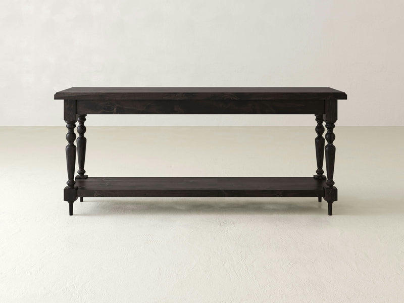 The Ella Sofa Table - Charred Ember by James+James is a dark wooden console table featuring a rectangular top and four turned legs, with an additional lower shelf connecting the legs near the base. The table is placed in an empty, light-colored room with a plain backdrop.