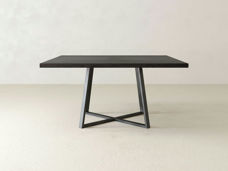 The Watson Square Dining Table - Charred Ember by James+James stands on a concrete floor against a light, plain wall. This minimalist wooden table with its dark finish features a modern design highlighted by an intersecting X-shaped geometric black metal base.