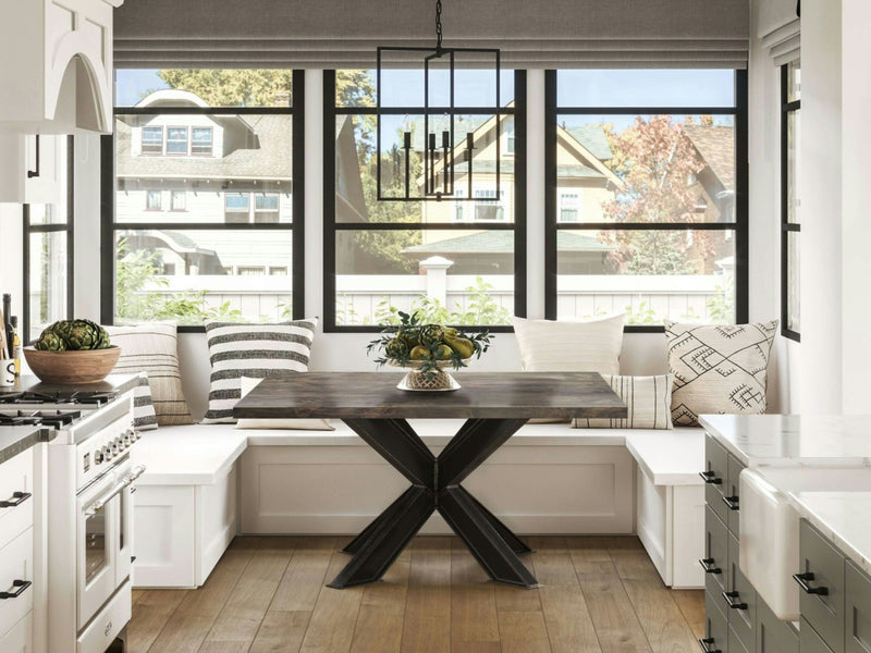 A cozy kitchen nook features the Shiloh Square Dining Table in Deep Grey with a black metal base, surrounded by white built-in bench seating with patterned cushions. Large windows offer a view of a suburban neighborhood. A geometric chandelier hangs overhead, while light wood flooring complements the space.