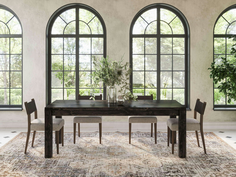 A dining room with a Jonathan Dining Table - Charred Ember from James+James, six gray upholstered chairs, and a decorative rug. The table features a vase with greenery and glassware. Arched windows in the background let in natural light, and there are potted plants on either side of the room.