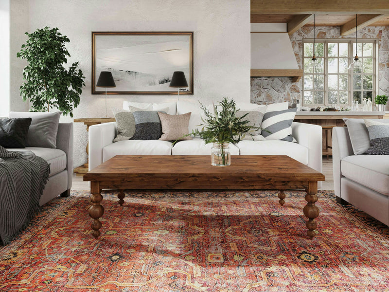 A cozy living room features two white sofas adorned with various pillows, a wooden Olivia Coffee Table - Tuscany by James+James holding a glass vase filled with greenery, a colorful patterned rug, and a framed landscape picture on the wall. In the background, you can see a rustic kitchen with wooden accents.