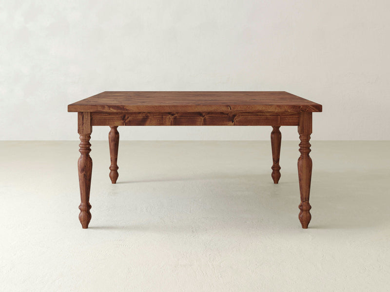 A square wooden dining table with a natural wood finish and four intricately carved legs, reminiscent of a French Country Square Dining Table from James+James, is positioned in a minimalist, light-colored room with a smooth floor and plain walls. The table's elegant design highlights its texture and grain.