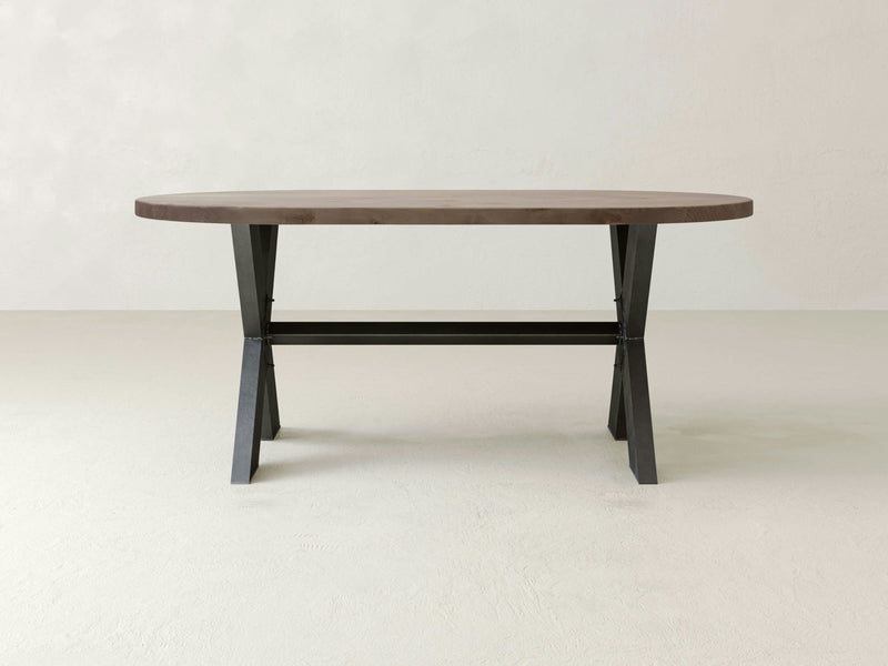 A James+James X-Base Oval Dining Table - Barn Wood with dark metal legs stands on a light-colored floor against a plain, off-white wall. The table features a simple, modern design with an x-frame support connecting the legs.