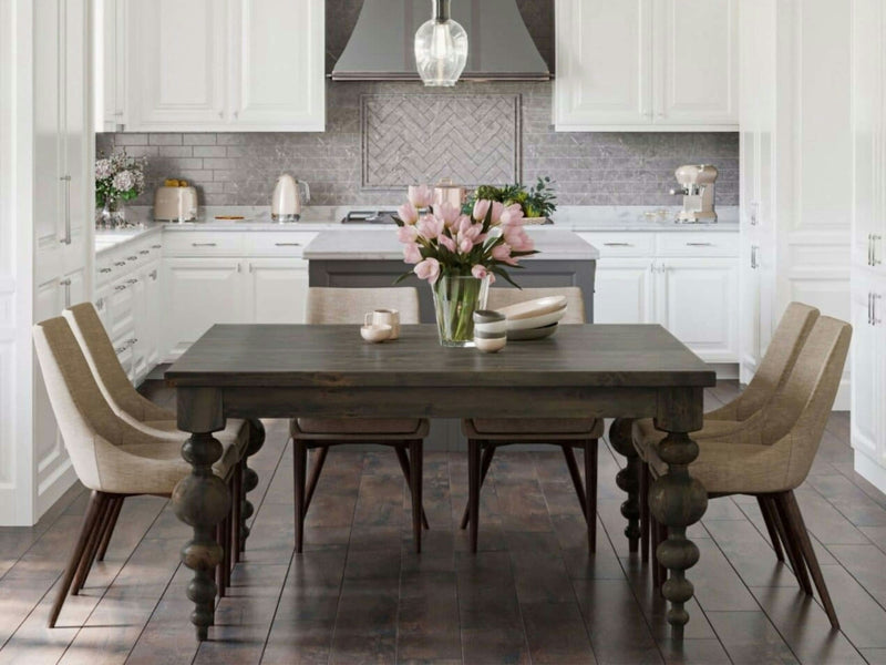 A modern kitchen with white cabinets, gray backsplash, and stainless steel appliances. Centered in the space is a James+James Olivia Square Dining Table in deep grey, surrounded by six light beige chairs. Atop the table are a vase of pink tulips, a bowl of fruit, and two cups. The floor features dark wood flooring.