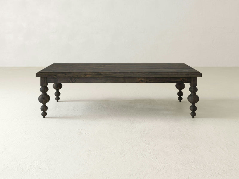 The Olivia Coffee Table - Deep Grey by James+James is a rectangular dark wooden table with four intricately carved legs, standing on a light-colored floor against a plain white background. This table boasts a minimalist design with a rustic appearance.