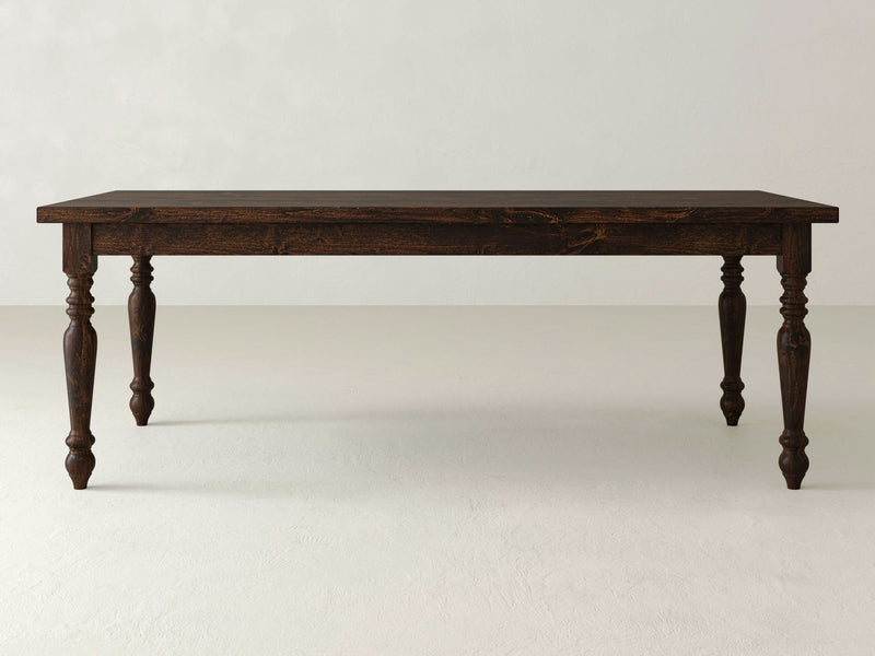 The French Country Dining Table - Tobacco by James+James stands against a plain, light-colored background. This dark wooden dining table features a rectangular top with beveled edges and intricately carved legs, showcasing detailed craftsmanship.