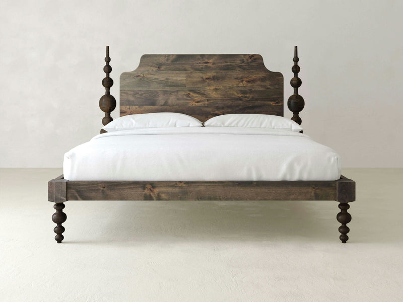 The Opal Bed by James+James features a high, carved headboard and intricately designed legs. The bed is crafted from dark-stained wood, adorned with white bedding and pillows, and set against a plain, light-colored backdrop.