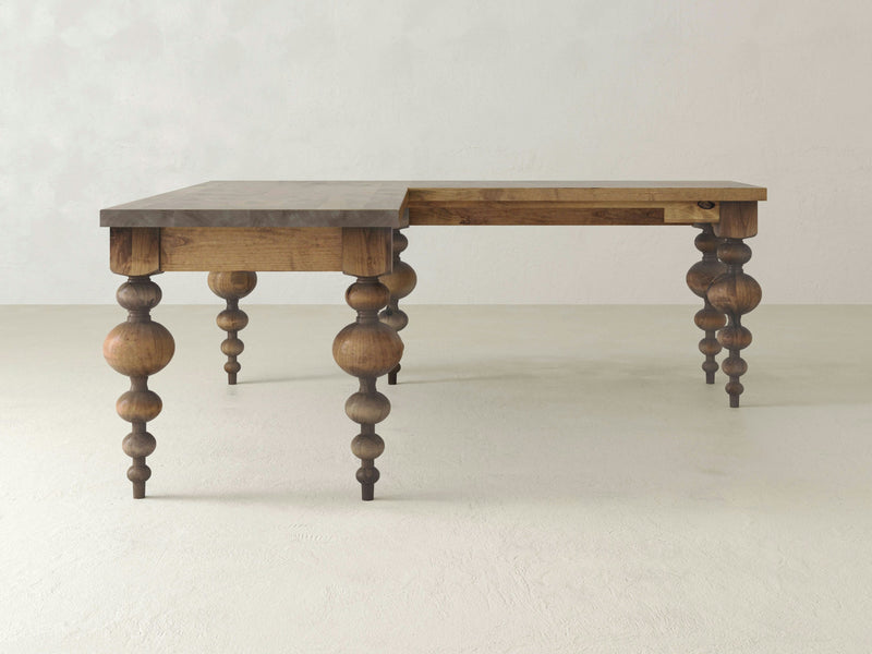 A detailed close-up of the Olivia Corner Desk - Harvest Wheat by James+James showcases its wooden surface, featuring a natural knot at the center and distinct grain patterns running horizontally across the light brown wood. The grain lines subtly curve around the knot, adding texture and depth to the image.