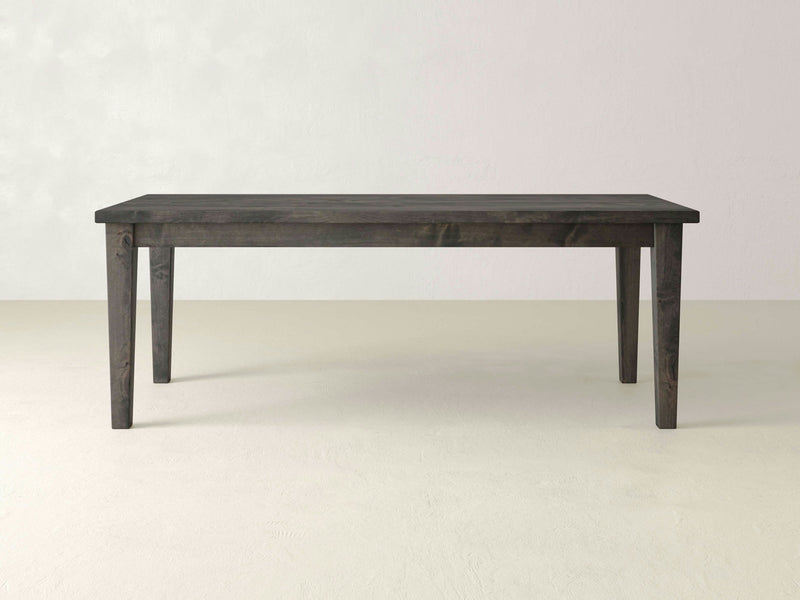 A rectangular Ava Parsons Dining Table in deep grey, crafted by James+James, stands gracefully in an empty room with light-colored walls and floor. Supported by four legs, the table features a dark, natural wood finish and exudes a simple, minimalist design.