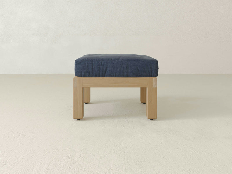 The Aria Outdoor Ottoman by James+James features a handcrafted wooden frame paired with a dark blue cushion. It stands against a plain, light-colored wall on a light gray floor, showcasing a minimalist design with clean lines and subtle hints of luxury that emphasize functionality.