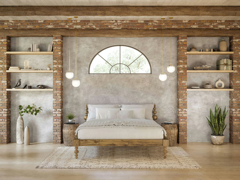 A cozy bedroom with a rustic, modern design features the Ophelia Bed in Harvest Wheat by James+James, adorned with white bedding and flanked by two wicker nightstands. Shelves built into the brick walls display decorative items. A large arched window is above the bed, and pendant lights hang from the wooden ceiling.