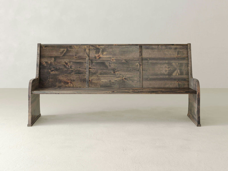 Introducing the Parish Pew Bench by James+James, a rustic wooden bench with a high backrest featuring three rectangular sections, a continuous seat, and two side supports. The bench boasts a deep grey finish with a weathered, natural wood look and is photographed against a plain, light-colored background.