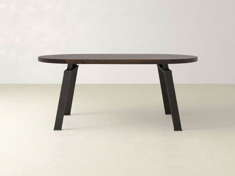 The Fulton Oval Dining Table - Tobacco by James+James features a sleek, minimalist design with an oval-shaped wooden top in a rich dark brown hue, complemented by black, angled legs. It stands on a light beige floor against a plain light grey wall, exuding contemporary elegance.