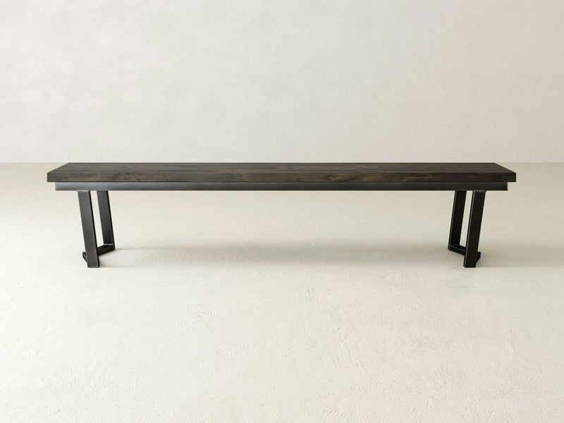 The James+James Arkwright Bench is a minimalist wooden bench crafted from solid hardwood with a dark finish, showcasing clean lines and sturdy black metal legs. The durable steel bases provide resilience, standing out against a plain, light-colored background.