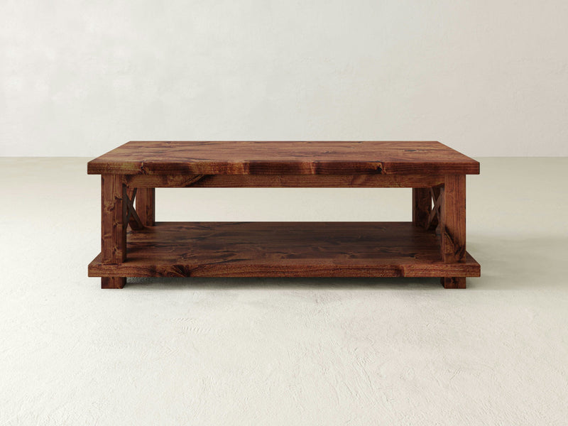 The Carter X Coffee Table - Tuscany by James+James is a rustic wooden piece featuring a rectangular top and a lower shelf for storage. It has a natural wood finish that beautifully highlights the grain and texture of the wood. The design includes sturdy, angular legs that support both the top and the shelf.