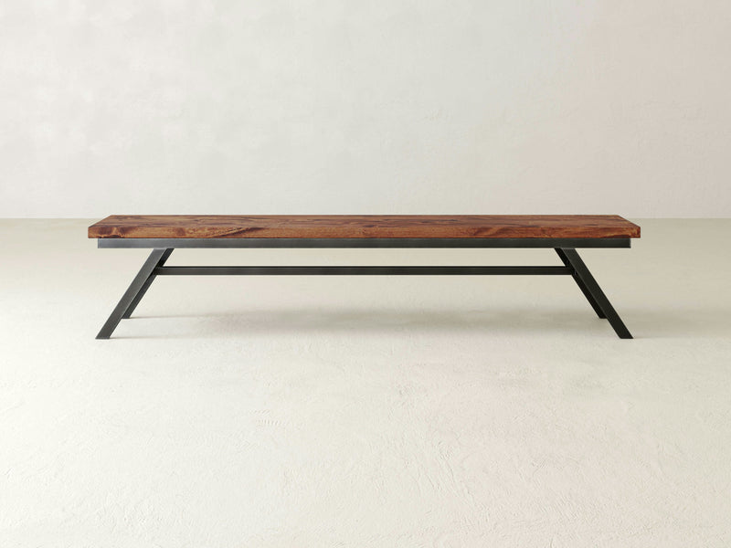 The Fulton Bench - Tuscany by James+James is a minimalist wooden bench featuring a rich, dark-brown seat complemented by sleek, black metal legs. Set against a light gray background, its design is modern and understated, emphasizing clean lines and simplicity.