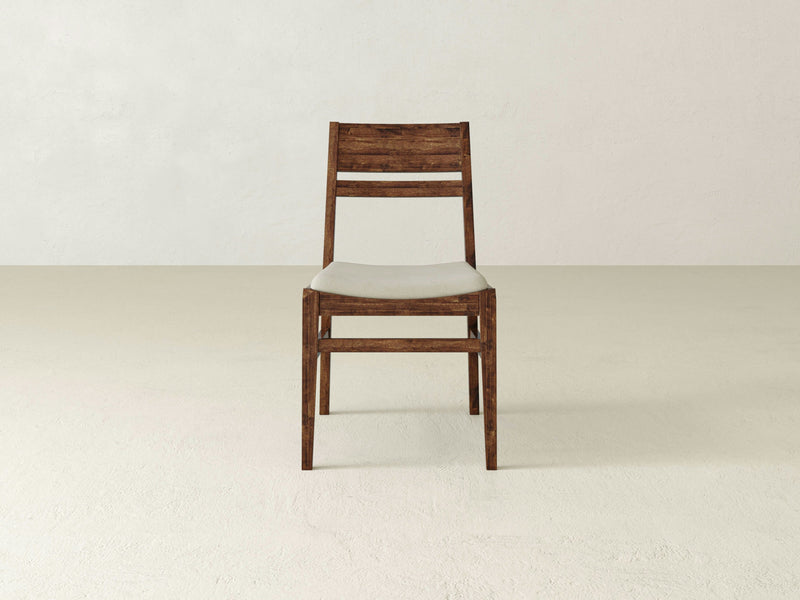 The Delilah Dining Chair - Tuscany by James+James graces a light-colored floor against a plain, off-white backdrop. This wooden chair features a slightly curved backrest and a white cushioned seat, embodying a simple, minimalist design.