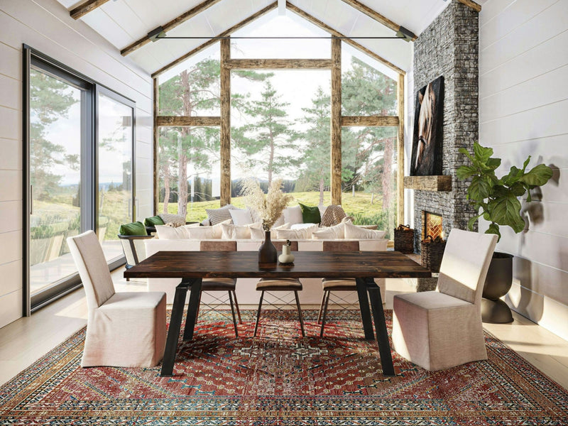 A spacious, modern living room with floor-to-ceiling windows offering a view of lush greenery. The room showcases the stunning Fulton Dining Table in Tobacco by James+James, paired with upholstered chairs, a stone fireplace, and a patterned rug. Natural light floods the space, enhancing its cozy ambiance.