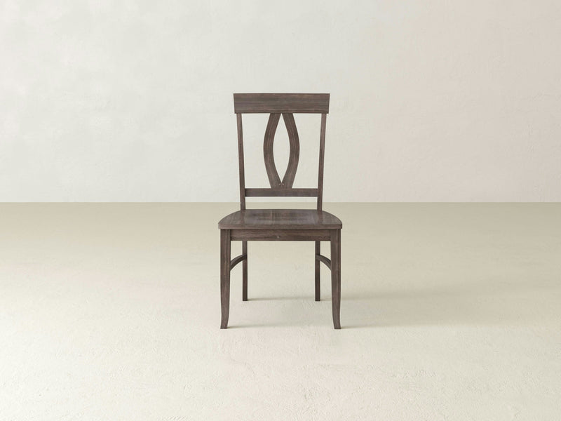 An Elizabeth Dining Chair - Barn Wood by James+James is placed in an empty room with light-colored walls and floor. The wooden chair, finished in a dark brown hue, features a rectangular backrest with a decorative cut-out pattern in the center and simple, straight legs.