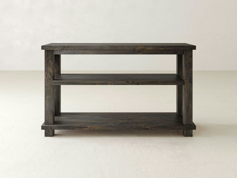 The Carter X Sofa Table - Deep Grey by James+James showcases a simple, rustic design. Featuring two lower shelves for additional storage, this dark wooden table stands out beautifully against a plain, light-colored background.