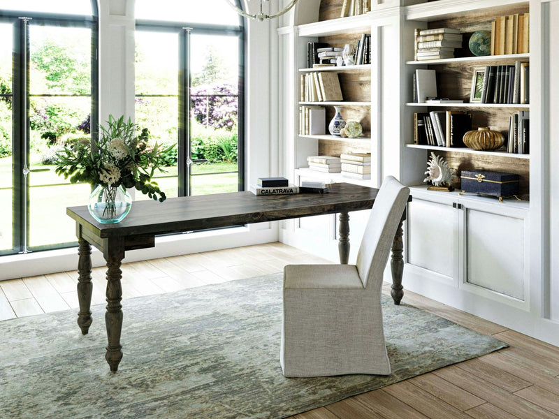 A bright home office with a large window revealing a lush garden. The room is accentuated by the Turned Leg Desk - Deep Grey from James+James, featuring a vase of flowers on top. A white chair and bookshelves filled with books and decor items add to the charm, while the light wooden floor is covered by a grayish-blue textured rug.