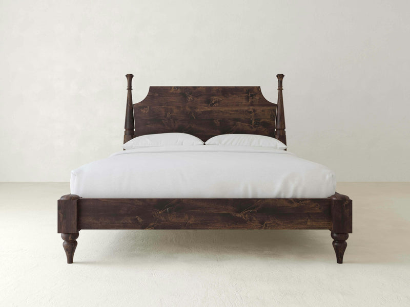 The James+James Stella Bed showcases superior craftsmanship with its dark wooden frame, tall carved posts on each corner, and a high, intricate headboard. The white mattress and pillows beautifully contrast against the light-colored wall, highlighting the bed's traditional elegance and timeless design.