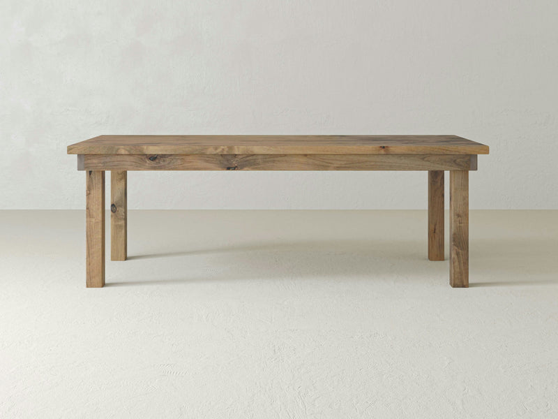 The James+James Farmhouse Dining Table features a minimalist design with a rectangular top and four straight legs, set against a plain, light-colored background. The visible wood grain adds rustic charm, emphasizing its sustainable hardwood construction.