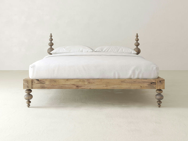 The Ophelia Bed by James+James, a minimalist wooden bed frame with intricately carved bedposts and a light finish, is the focal point of a plain, light-colored room. The elegant bedroom furniture is complemented by crisp white bedding, including pillows and a neatly spread blanket. The overall aesthetic is simple and sophisticated.