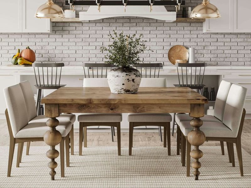 A modern kitchen features the Olivia Square Dining Table in Harvest Wheat by James+James, accompanied by six chairs upholstered in light gray fabric. A potted plant sits as a centerpiece on the table. The background includes white cabinets, a white brick wall, and stainless steel appliances.