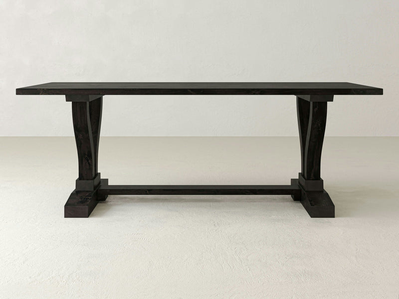 A Vera Dining Table - Charred Ember by James+James, featuring a rectangular dark wooden top and two thick, curved legs connected by a horizontal support bar, stands on a white textured floor against a plain light grey wall. The design is simple and sturdy.