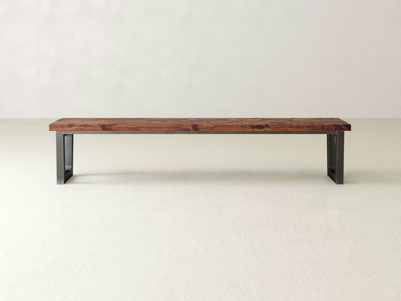 A minimalist James+James Trapezoid Bench - Tuscany features a rectangular dark wood top and black metal legs, placed against a plain light-colored wall and floor background. The design is simple and modern.