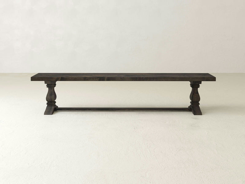 The Heirloom Bench - Deep Grey from James+James is a dark wooden bench featuring a flat rectangular seat and ornate, turned legs. It includes a connecting stretcher for added support and stability. The bench is set against a plain off-white wall with a light-colored floor in the background.