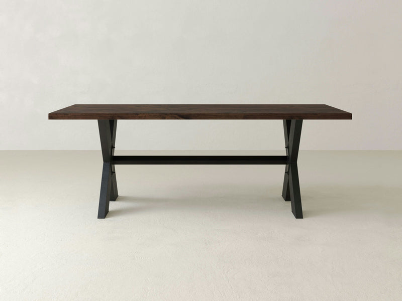 The X-Base Dining Table - Tobacco by James+James is positioned against a light, neutral background. This minimalist rectangular table features a dark brown tabletop and black trestle legs, showcasing a clean, simple design that highlights its sturdy construction and modern aesthetic.