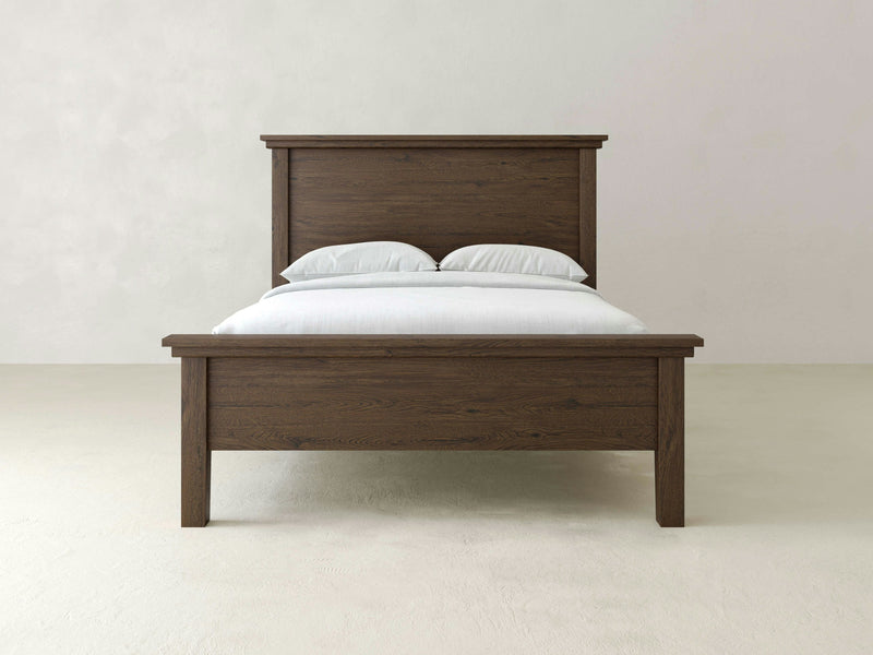 An Ivy Bed - Charred Ember by James+James, with a simple, rustic design, is placed against a plain white wall. The bed features a high headboard and a matching footboard, adorned with neatly arranged white bed linens and pillows. The wood boasts a rich, dark brown finish.