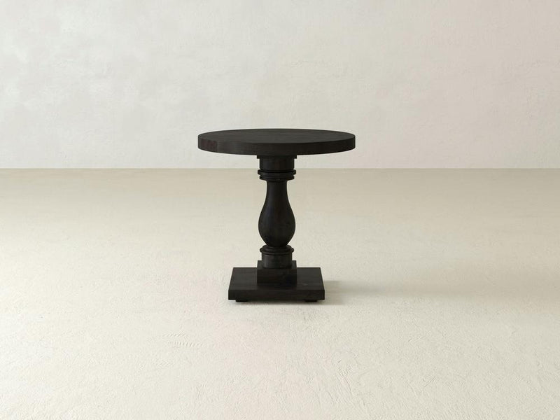 The Vivien Side Table - Charred Ember by James+James features a dark wooden circular top resting on an ornately carved, sturdy single leg, all supported by a square base. The table is showcased against a plain, light-colored background.