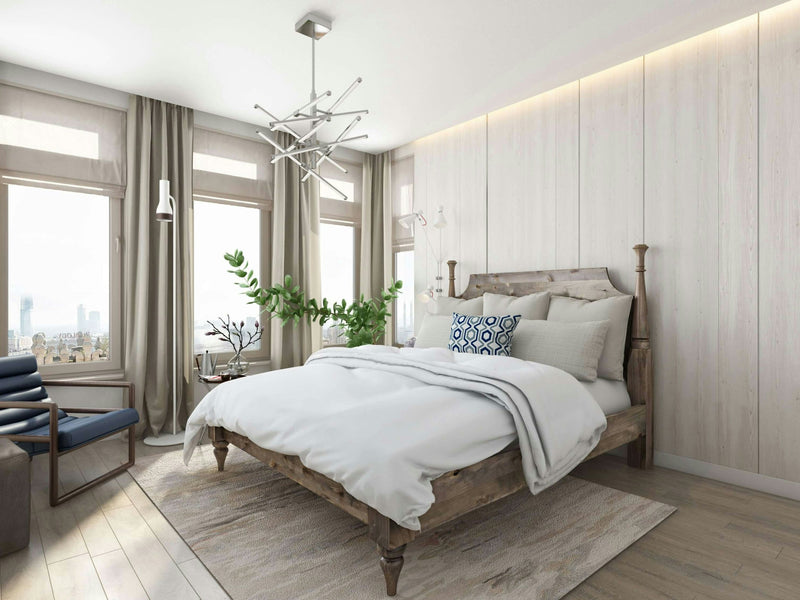 A modern bedroom features the Stella Bed made from barn wood by James+James, adorned with white linens and patterned pillows. Large windows with light curtains allow natural light to flood the room. The space includes a contemporary chandelier, indoor plants, and a lounge chair.