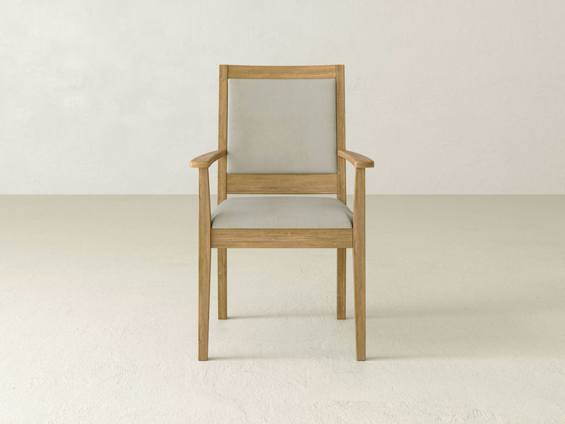 The Gina Arm Dining Chair - Harvest Wheat by James+James, featuring a light beige cushioned seat and backrest, is centered in a minimalist room with light-colored walls and floor. The chair boasts simple, clean lines and wooden armrests.