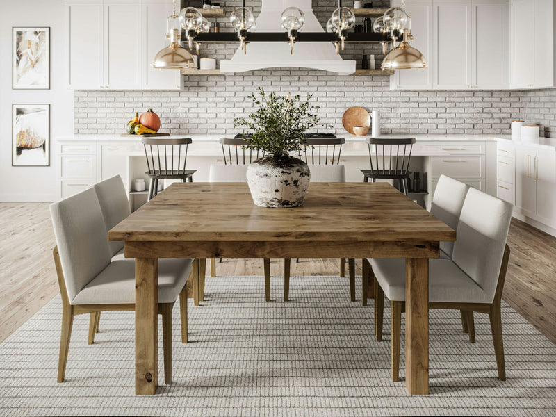 A modern dining room features the Farmhouse Square Dining Table - Harvest Wheat by James+James, complemented by grey upholstered chairs. A large potted plant sits in the center of the table. The background includes a kitchen with white cabinetry, a white brick backsplash, and pendant lights hanging above a kitchen island.