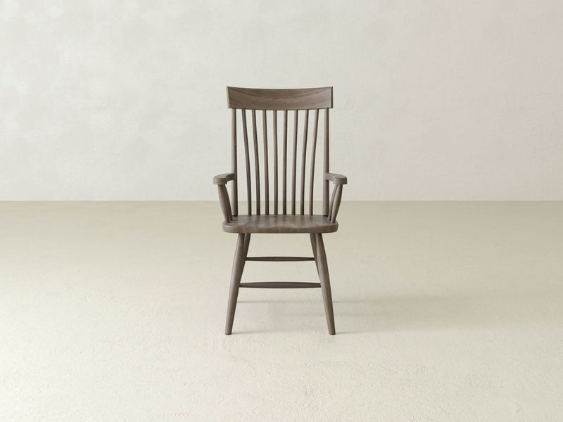 The Eleanor Arm Dining Chair - Barn Wood by James+James, a single wooden chair with a high back and vertical slats, featuring armrests and a slightly curved seat, is positioned against a plain, light-colored wall and sits on a smooth, light-colored floor. The design is simple and modern.