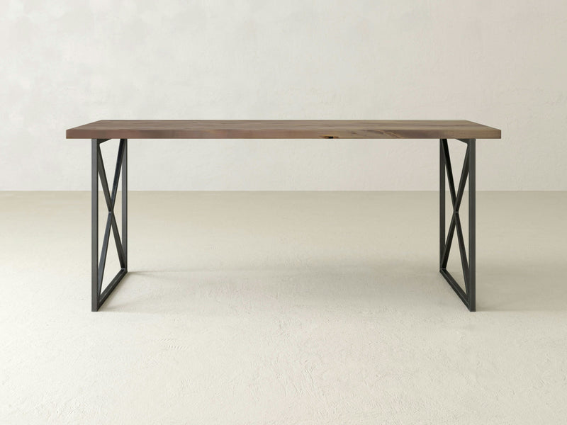 The Luca Desk - Barn Wood by James+James features a minimalist design with a light brown rectangular wooden top and black metal legs forming an "X" shape on each side, set against a plain white background. The overall look is simple and modern.