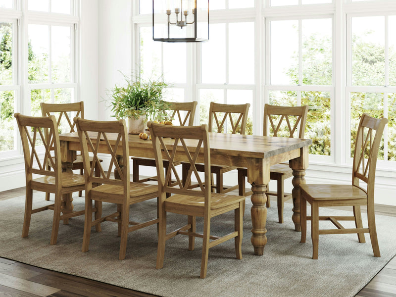 A spacious dining room featuring a large James+James Baluster Dining Table in Harvest Wheat, surrounded by eight matching wooden chairs with X-back designs. The table is adorned with a potted plant and small decorative items. The room is well-lit by natural light streaming through large windows.