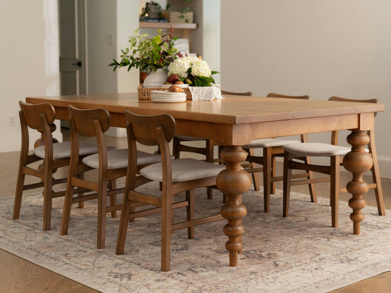 An Olivia Dining Table in Harvest Wheat by James+James with ornate, turned legs is surrounded by six matching chairs with upholstered seats. A floral centerpiece and a wooden bowl adorn the table. The setup is placed on a patterned area rug in a bright, airy room with a light color palette.