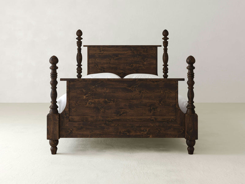 The Catherine Bed in Tobacco by James+James features a dark wooden frame with an intricate design and four tall, rounded posts at each corner. The solid headboard and footboard showcase visible wood grain, contributing to its rustic appearance. This bed is placed against a white wall on a light floor.