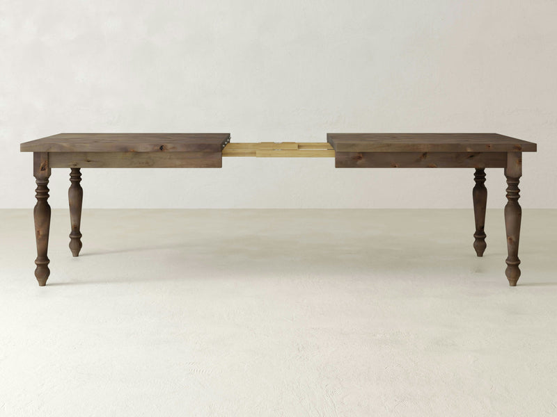 The James+James French Country Expandable Dining Table - Barn Wood, featuring ornate turned legs, is displayed in an extended position with its central mechanism exposed and ready for an additional leaf insert. The table is set against a plain, off-white background.