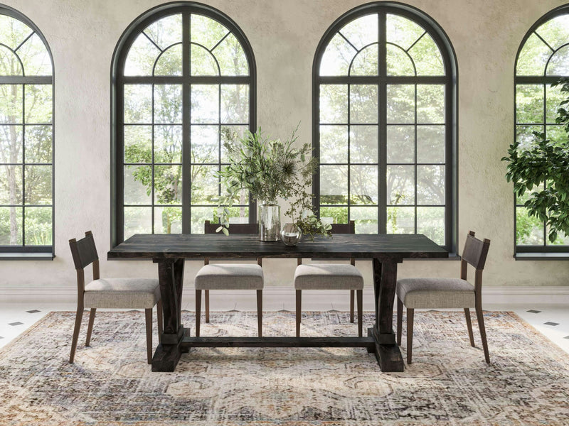 A dining room with large, arched windows providing natural light features the Vera Expandable Dining Table in Charred Ember from James+James. The table is adorned with green plants and two transparent vases. Surrounding the table are four wooden chairs with light gray upholstered seats, and a patterned rug lays on the floor beneath them.