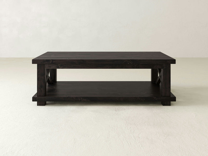 The Carter X Coffee Table by James+James features a dark wooden rectangular top and a lower shelf, exuding rustic elegance against a plain, light-colored background. Its simple and sturdy design with straight corner legs perfectly complements any contemporary farmhouse decor.