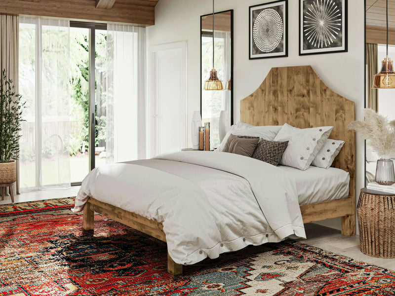 A cozy bedroom featuring the Megan Bed in Harvest Wheat from James+James, adorned with a white comforter and an assortment of pillows. Decorative artworks are hung above the bed, while natural light streams in from a large window draped with sheer curtains. The floor is covered by a vibrant, multicolored rug.