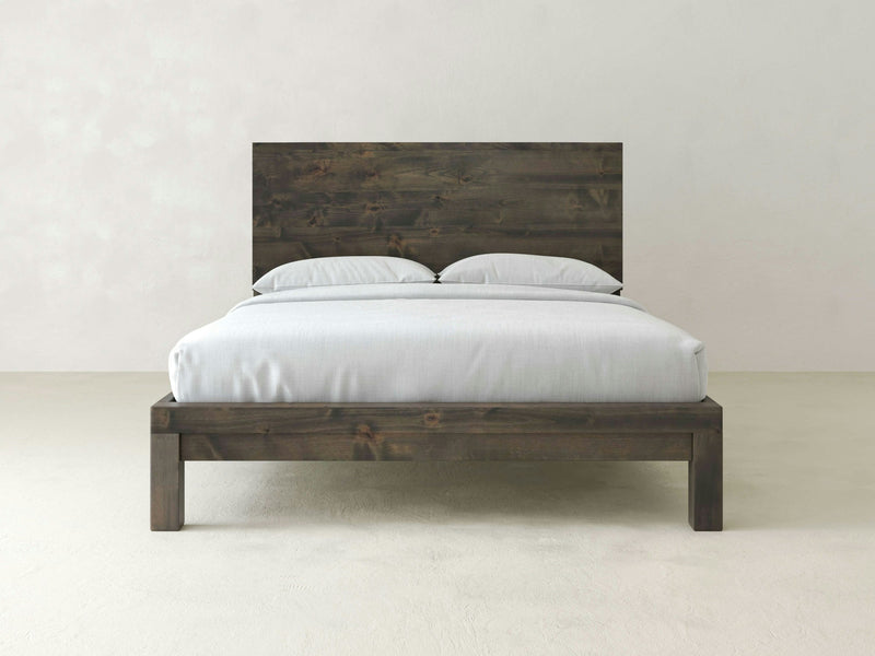 The Sawyer Bed - Deep Grey from James+James, featuring a minimalist dark wooden frame and headboard, is topped with a white mattress and two matching white pillows. It is centered against a light gray wall in an otherwise empty room, creating a simple and modern aesthetic.