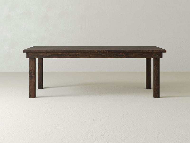 A rectangular, dark brown wooden Farmhouse Dining Table - Tobacco by James+James, featuring four straight legs and a simple design with clean lines and no decorative elements, stands against a pale green, textured wall on a light-colored floor.